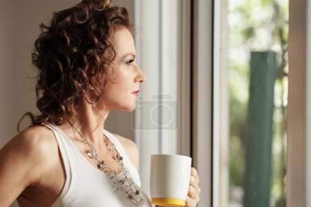 Foto de Pensativo mujer madura grave beber taza de café y mirando fuera a través de la ventana del apartamento - Imagen libre de derechos