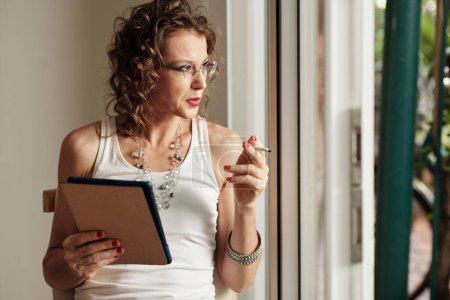 Foto de Mujer madura en camiseta sin mangas y gafas sosteniendo la tableta, fumando y mirando por la ventana - Imagen libre de derechos