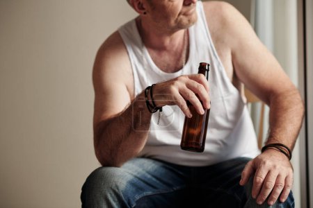 Téléchargez les photos : Image recadrée d'un homme mûr en maillot blanc buvant de la bière et regardant dehors par la fenêtre - en image libre de droit