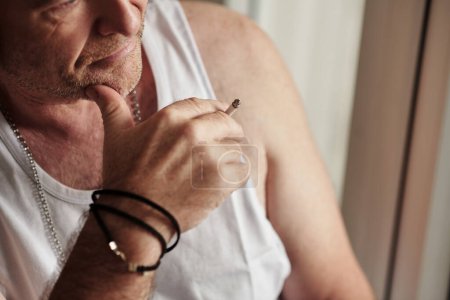 Téléchargez les photos : Pensive homme mûr en blanc maillot de corps fumer cigarette - en image libre de droit