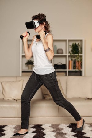 Foto de Mujer madura glamurosa que usa auriculares VR cuando hace ejercicio con pesas - Imagen libre de derechos