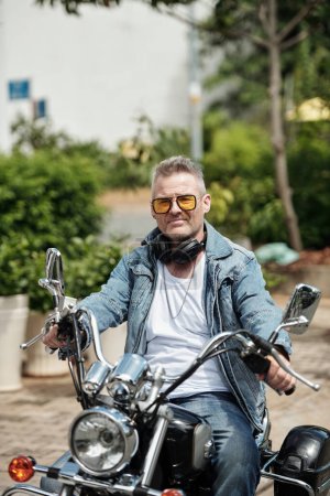 Photo for Portrait of cool mature man in yellow glasses and demin jacket sitting on motorbike - Royalty Free Image