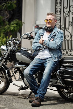Foto de Retrato de hombre maduro confiado en jeans y chaqueta de demonio apoyado en la motocicleta cuando espera a su novia - Imagen libre de derechos