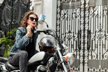 Foto de Elegante mujer madura glamorosa comprobando su cabello y maquillaje en el espejo de la motocicleta antes de montar - Imagen libre de derechos