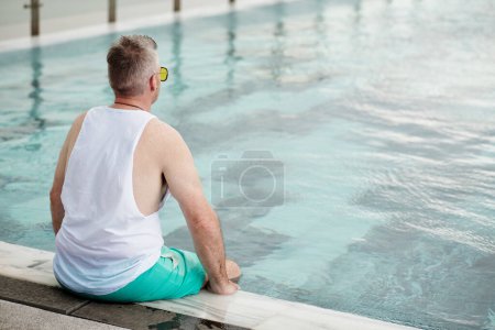 Photo for Pensive mature man sitting on swimming pool edge and looking at water, view from back - Royalty Free Image
