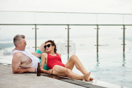 Téléchargez les photos : Élégant couple d'âge mûr se détendre et boire de la bière par piscine - en image libre de droit