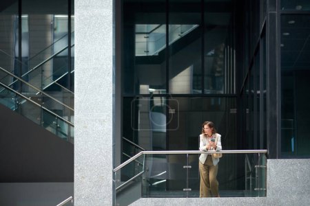 Téléchargez les photos : Femme d'affaires pensive avec smartphone s'appuyant sur la rampe et détournant les yeux - en image libre de droit