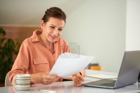 Téléchargez les photos : Souriante femme d'affaires mature lisant un rapport imprimé avec des données financières - en image libre de droit