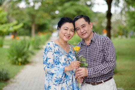 Photo for Portrait of happy senior Vietnamese couple hugging and holding rose flower when standing on path in city park - Royalty Free Image