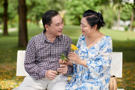Téléchargez les photos : Vietnamien senior homme donnant rose jaune à petite amie quand ils sont assis sur le banc dans le parc - en image libre de droit