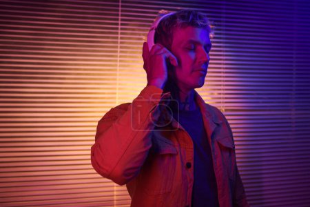 Téléchargez les photos : Portrait de jeune homme debout dans la lumière du néon et à l'écoute de sa piste musicale préférée dans les écouteurs - en image libre de droit