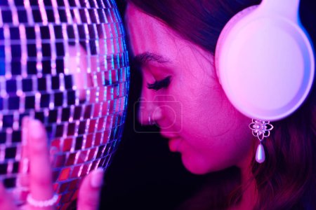 Foto de Mujer joven escuchando música en auriculares y apoyando la frente en la bola disco - Imagen libre de derechos