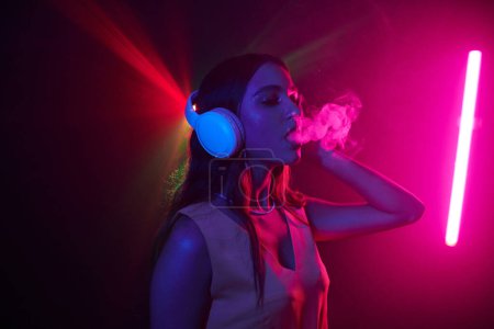 Téléchargez les photos : Jeune femme jouissant de la vie nocturne, elle danse dans des écouteurs et fume - en image libre de droit