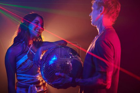 Foto de Sonriente bailarina del club nocturno coqueteando con un joven sosteniendo una bola de discoteca - Imagen libre de derechos