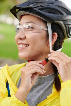 Photo for Happy female cyclist putting on bicycle helmet to ride in park - Royalty Free Image