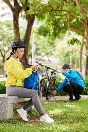 Téléchargez les photos : Femme prenant de l'eau vitaminée du sac à dos lorsque son petit ami ajustant le support du pied de vélo - en image libre de droit
