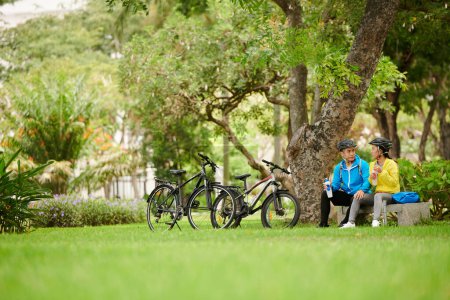 Photo for Serious young Asian couple drinking water under tree in park after bicycle ride - Royalty Free Image