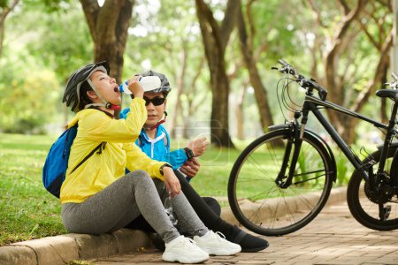 Photo for Couple tired after cycling sitting on pavement, drinking water and talking - Royalty Free Image