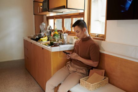 Foto de Joven sentado en el banco en la cocina y probando amigos en el teléfono inteligente - Imagen libre de derechos