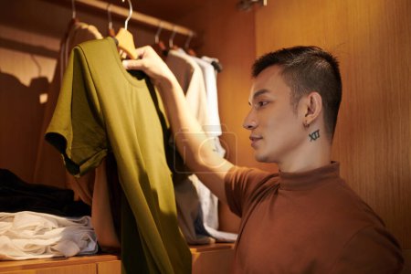 Photo for Young man standing in closet and holding hanger with t-short when choosing what to wear today - Royalty Free Image