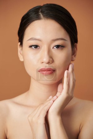 Téléchargez les photos : Portrait d'une jeune femme sérieuse avec un maquillage naturel touchant le visage et regardant la caméra - en image libre de droit