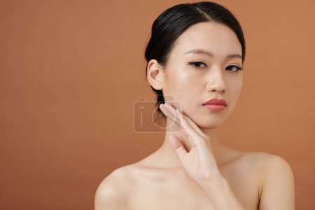 Photo for Portrait of serious young woman touching face and looking at camera, isolated on brown background - Royalty Free Image