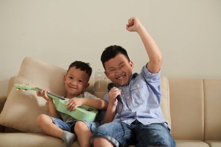 Photo for Excited boy singing song into microphone when his little brother playing ukulele at home - Royalty Free Image