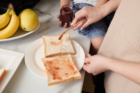 Foto de Las manos de la madre extendiendo mermelada de bayas en rebanada de pan al cocinar el desayuno - Imagen libre de derechos
