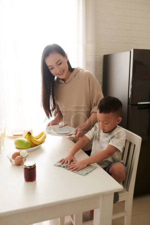 Téléchargez les photos : Petit garçon souriant essuyant table de cuisine avec un chiffon doux après le petit déjeuner lorsque la mère collecte des plats - en image libre de droit