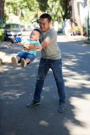 Foto de Alegre padre jugando con pequeño hijo en la carretera en frente de la casa - Imagen libre de derechos