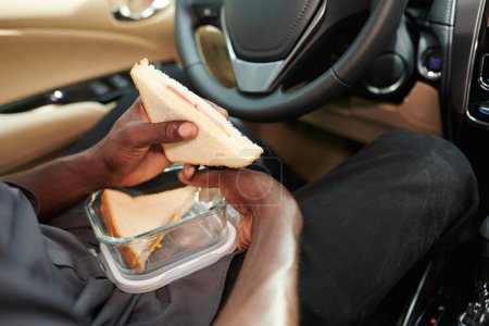 Téléchargez les photos : Mains de chauffeur de taxi manger savoureux sandwich maison qu'il a fait à la maison pour le déjeuner - en image libre de droit