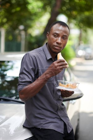 Téléchargez les photos : Portrait du chauffeur de taxi penché sur la voiture et manger un sandwich pour le déjeuner - en image libre de droit