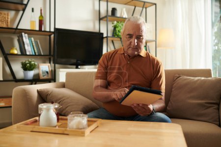 Téléchargez les photos : Homme âgé boire du thé et lire l'article de nouvelles sur tablette informatique à la maison - en image libre de droit