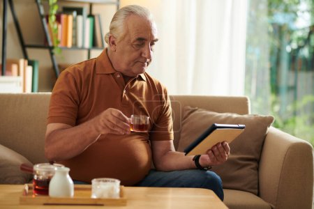 Photo for Senior man drinking tea and watching news on tablet computer in the morning - Royalty Free Image
