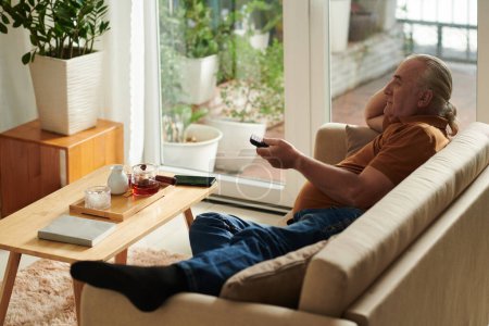 Téléchargez les photos : Homme âgé se détendre sur le canapé à la maison et regarder un nouveau spectacle ou un film à la télévision - en image libre de droit