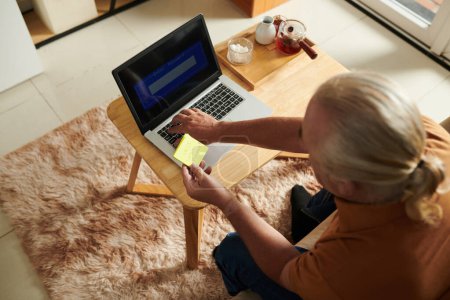 Téléchargez les photos : Senior homme entrant mot de passe écrit sur la note collante lors de la connexion au site Web - en image libre de droit