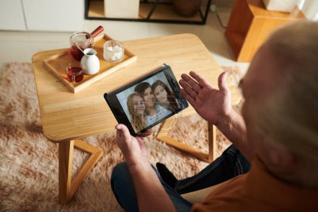 Téléchargez les photos : Homme âgé avec tablette vidéo appelant sa fille et ses petites-filles lors de la consommation de thé à la maison - en image libre de droit