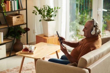 Téléchargez les photos : Homme âgé dans les écouteurs regarder des vidéos de musique sur tablette - en image libre de droit