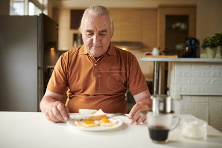 Foto de Hombre mayor comiendo huevos fritos con pan y café para desayunar en casa - Imagen libre de derechos