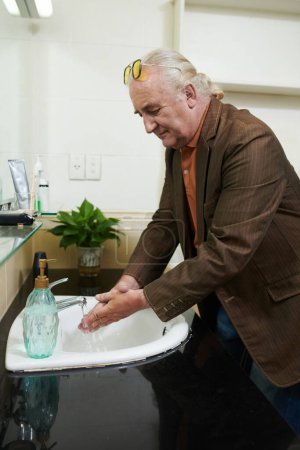 Téléchargez les photos : Homme âgé avec queue de cheval se laver les mains à fond dans la salle de bain à la maison - en image libre de droit
