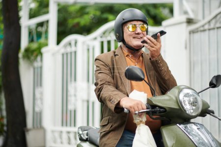 Téléchargez les photos : Joyeux vieil homme assis sur scooter et enregistrement message vocal pour ami ou petite amie - en image libre de droit