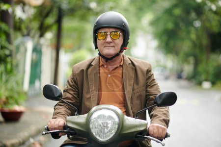 Téléchargez les photos : Homme âgé souriant en lunettes jaunes et casque de protection à cheval sur scooter - en image libre de droit