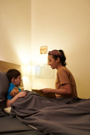 Téléchargez les photos : Mère souriante lisant le livre pour son fils préadolescent couché au lit avec un ours en peluche - en image libre de droit