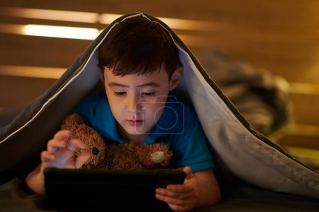 Téléchargez les photos : Garçon avec ours en peluche couché sur le lit et jouer au jeu sur tablette numérique la nuit - en image libre de droit