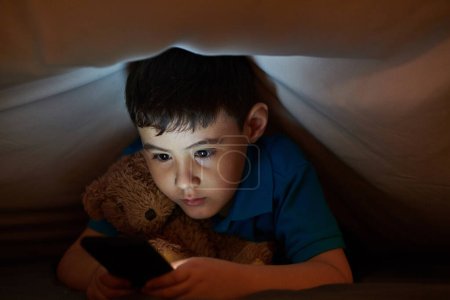Foto de Niño abrazando oso de peluche al jugar juego en el teléfono inteligente bajo la manta en la noche - Imagen libre de derechos