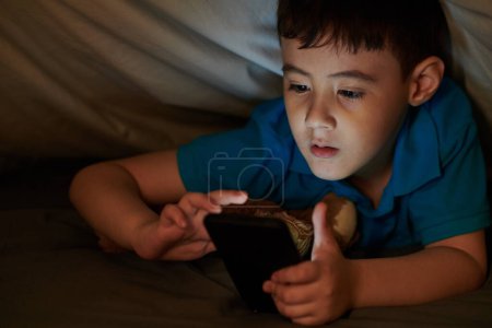 Photo for Child watching videos or playing games on smartphone under blanket at night - Royalty Free Image
