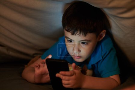 Téléchargez les photos : Preteen garçon couché sous couverture et surfer sur les médias sociaux sur smartphone tard dans la nuit - en image libre de droit
