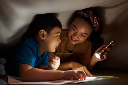 Foto de Sonriente madre e hijo leyendo libro bajo manta hasta altas horas de la noche - Imagen libre de derechos