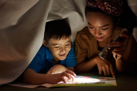 Téléchargez les photos : Mère lampe de poche brillante sur la page du livre lorsque son fils lit une histoire effrayante - en image libre de droit
