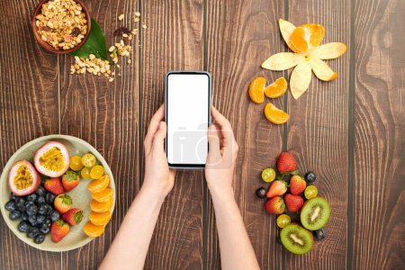 Foto de Manos sosteniendo teléfono inteligente sobre mesa de madera con frutas, bayas y tazón de muesli - Imagen libre de derechos